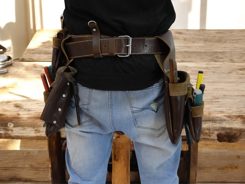 May include: A brown leather tool belt with multiple pockets and pouches, worn over blue jeans. The belt has a metal buckle and is secured with a brown leather strap.