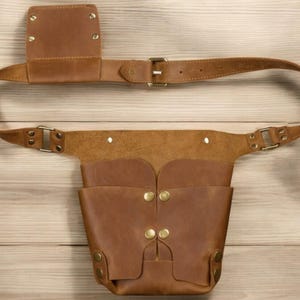 Pu&ograve; includere: Una cintura portautensili in pelle marrone con una tasca grande e una tasca pi&ugrave; piccola sul lato. La cintura ha una fibbia in ottone ed &egrave; fissata con rivetti in ottone.