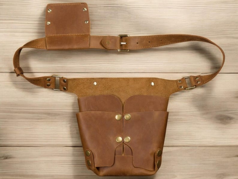 Pu&ograve; includere: Una cintura portautensili in pelle marrone con una tasca grande e una tasca pi&ugrave; piccola sul lato. La cintura ha una fibbia in ottone ed &egrave; fissata con rivetti in ottone.
