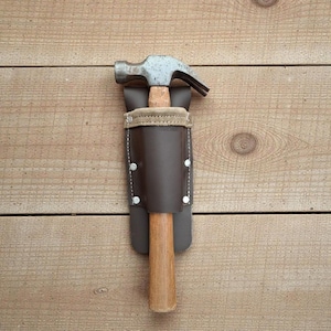 May include: A claw hammer with a wooden handle is secured in a brown leather tool holder. The holder is stitched with white thread and secured with silver rivets. The hammer head is made of metal.