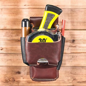 May include: A brown leather tool belt with a 30-foot tape measure, a pencil, and a cylindrical tool. The tape measure has a yellow and black casing. The belt is attached to a wooden surface.