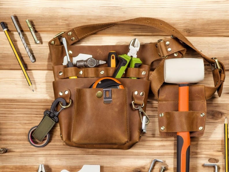Pu&ograve; includere: Una cintura portautensili in pelle marrone con pi&ugrave; tasche e una variet&agrave; di strumenti, tra cui un martello, una pinza, un metro a nastro e un mazzuolo. La cintura &egrave; appesa su una superficie di legno.