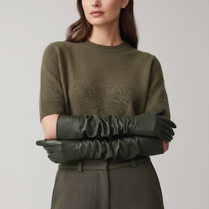 May include: Olive green leather gloves with a ruched design. The gloves are long, extending past the elbows. The model is wearing a matching olive green sweater and pants. The gloves are a stylish accessory.