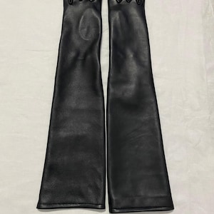 May include: A pair of long, black leather gloves. The gloves are sleek and reach well past the elbows. The leather appears smooth and the gloves are laid out on a white surface.