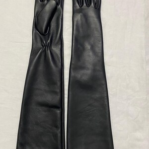 May include: A pair of long, black leather gloves. The gloves are sleek and form-fitting, extending well past the wrist. The leather appears smooth and has a slight sheen. The gloves are laid out side by side on a white surface.