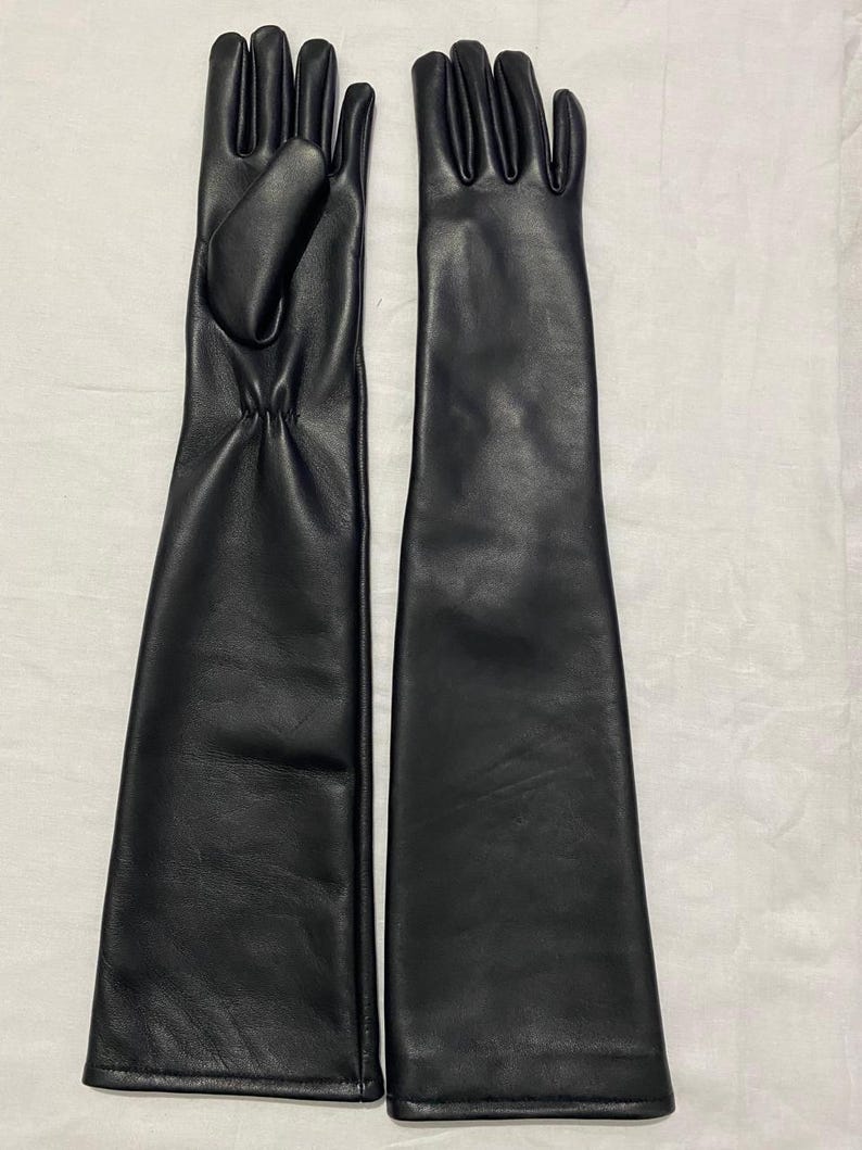 May include: A pair of long, black leather gloves. The gloves are sleek and form-fitting, extending well past the wrist. The leather appears smooth and has a slight sheen. The gloves are laid out side by side on a white surface.