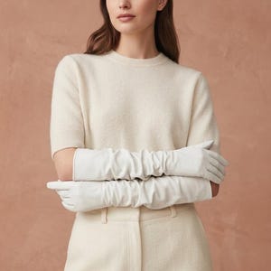 May include: A pair of long, white leather gloves featuring a ruched, gathered texture that extends past the elbows. The gloves are worn with a short-sleeved, cream-colored knit sweater and matching cream-colored trousers with a visible waistband.