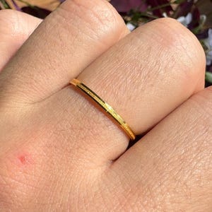 May include: A thin, gold ring with a textured surface. The ring is a simple band design, and is shown on a hand. The background features purple leaves and sunlight.