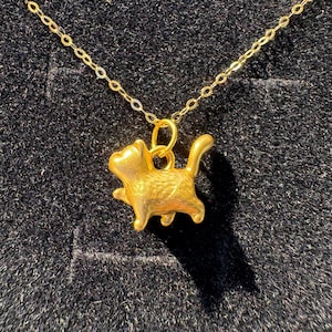 May include: A gold-coloured necklace with a cat-shaped pendant. The pendant shows a detailed cat with paws and a tail. The fine gold chain hangs against a dark background. The pendant is approximately 2 cm in length.