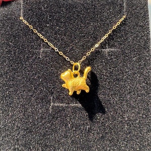 May include: A gold-colored necklace featuring a small, three-dimensional cat pendant in a walking pose. The pendant is suspended from a delicate gold-colored chain, showcasing a smooth, metallic finish with detailed cat features.