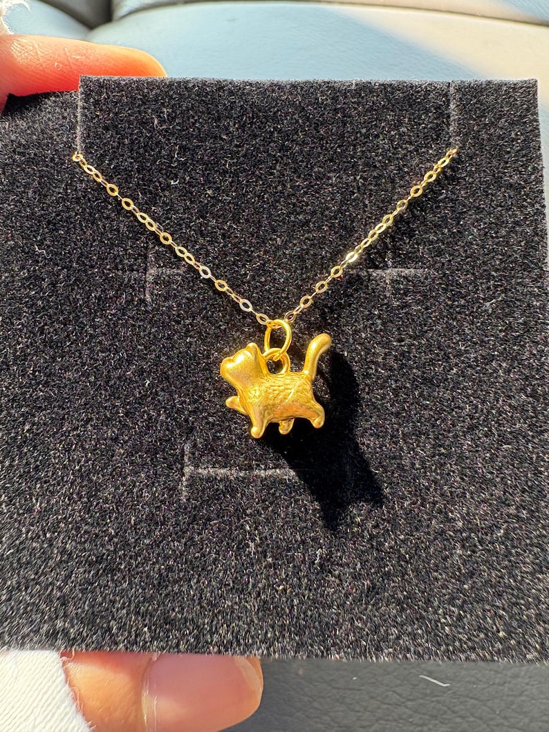 May include: A gold-colored necklace featuring a small, three-dimensional cat pendant in a walking pose. The pendant is suspended from a delicate gold-colored chain, showcasing a smooth, metallic finish with detailed cat features.