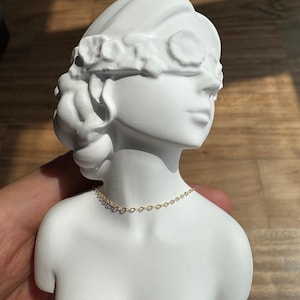 May include: A white matte bust sculpture of a figure with a floral headband covering the eyes, featuring a delicate gold-toned chain necklace resting around the neck. The sculpture is smooth and solid, depicting the head and shoulders of a person.