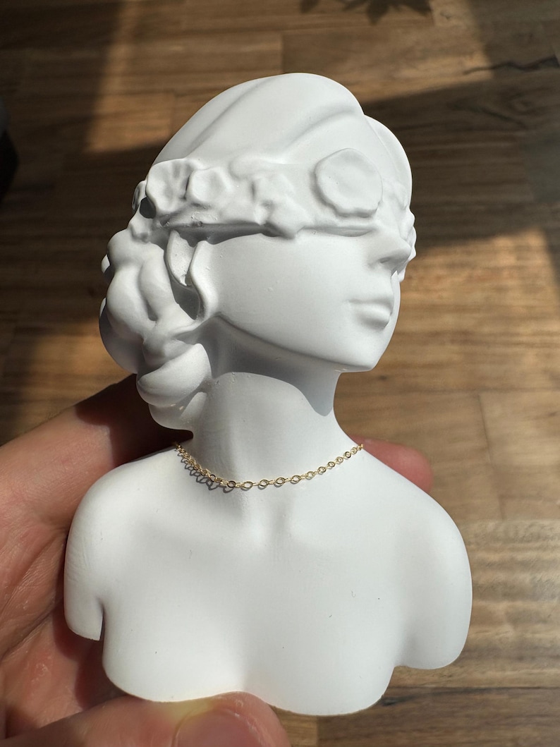 May include: A white matte bust sculpture of a figure with a floral headband covering the eyes, featuring a delicate gold-toned chain necklace resting around the neck. The sculpture is smooth and solid, depicting the head and shoulders of a person.