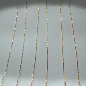 May include: Six delicate gold chains arranged vertically in parallel lines. Each chain features a series of small, reflective, oval-shaped links that catch the light, creating a shimmering effect across the length of the thin, metallic jewelry strands.
