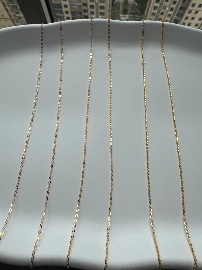 May include: Six delicate gold chains arranged vertically in parallel lines. Each chain features a series of small, reflective, oval-shaped links that catch the light, creating a shimmering effect across the length of the thin, metallic jewelry strands.