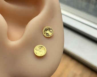 May include: Two small, round, gold-colored stud earrings featuring a textured surface with a pattern of overlapping circles. The earrings have a thin, raised border around the edge and are displayed on a neutral-toned ear model.