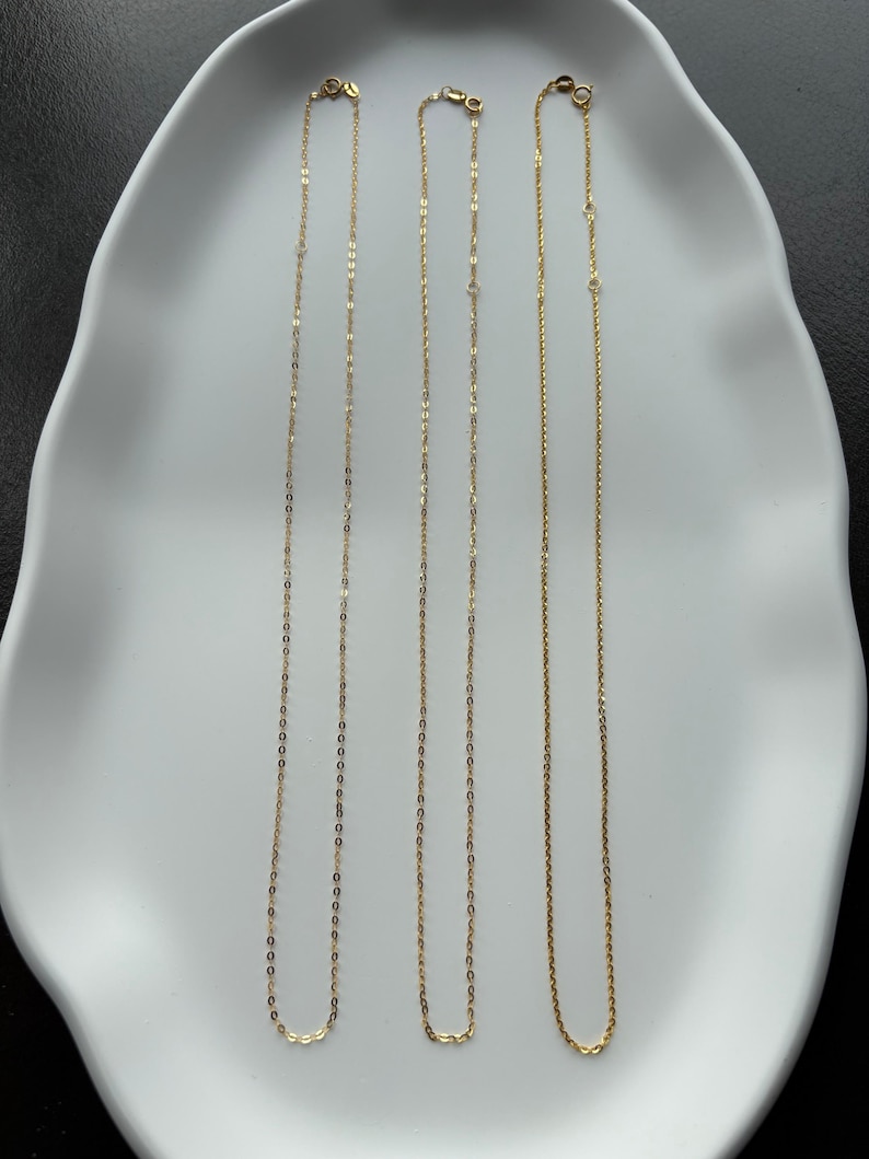 May include: Three delicate gold-toned necklaces with fine link chains, each featuring a small circular clasp at the top. The necklaces are arranged vertically, displaying a consistent metallic finish and a simple, slender design structure.