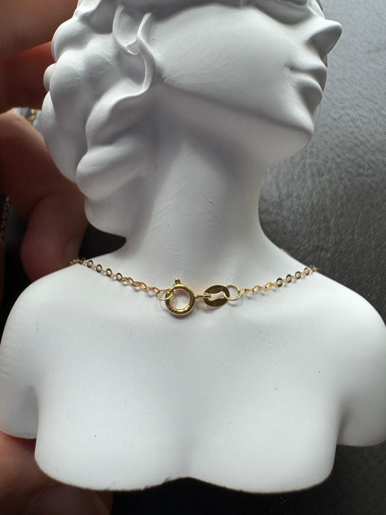 May include: A delicate gold chain necklace featuring a round spring ring clasp and a small oval tag. The chain is composed of thin, interlocking circular links, showcasing a polished metallic finish that rests against a white sculpted bust.