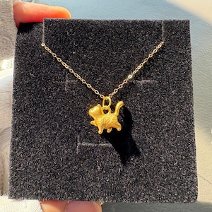 May include: A gold-toned necklace featuring a small, three-dimensional cat pendant in a walking pose. The pendant is suspended from a delicate, thin gold-toned chain. The jewelry is displayed against a square, black, textured velvet-like backing.