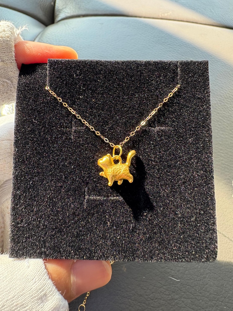 May include: A gold-toned necklace featuring a small, three-dimensional cat pendant in a walking pose. The pendant is suspended from a delicate, thin gold-toned chain. The jewelry is displayed against a square, black, textured velvet-like backing.