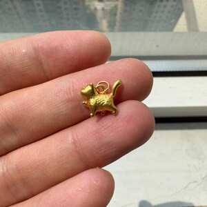 May include: A small, gold-colored metal charm shaped like a stylized cat in a walking pose. The charm features a textured surface and includes a small circular jump ring attached to the top of the cat's back for hanging purposes.