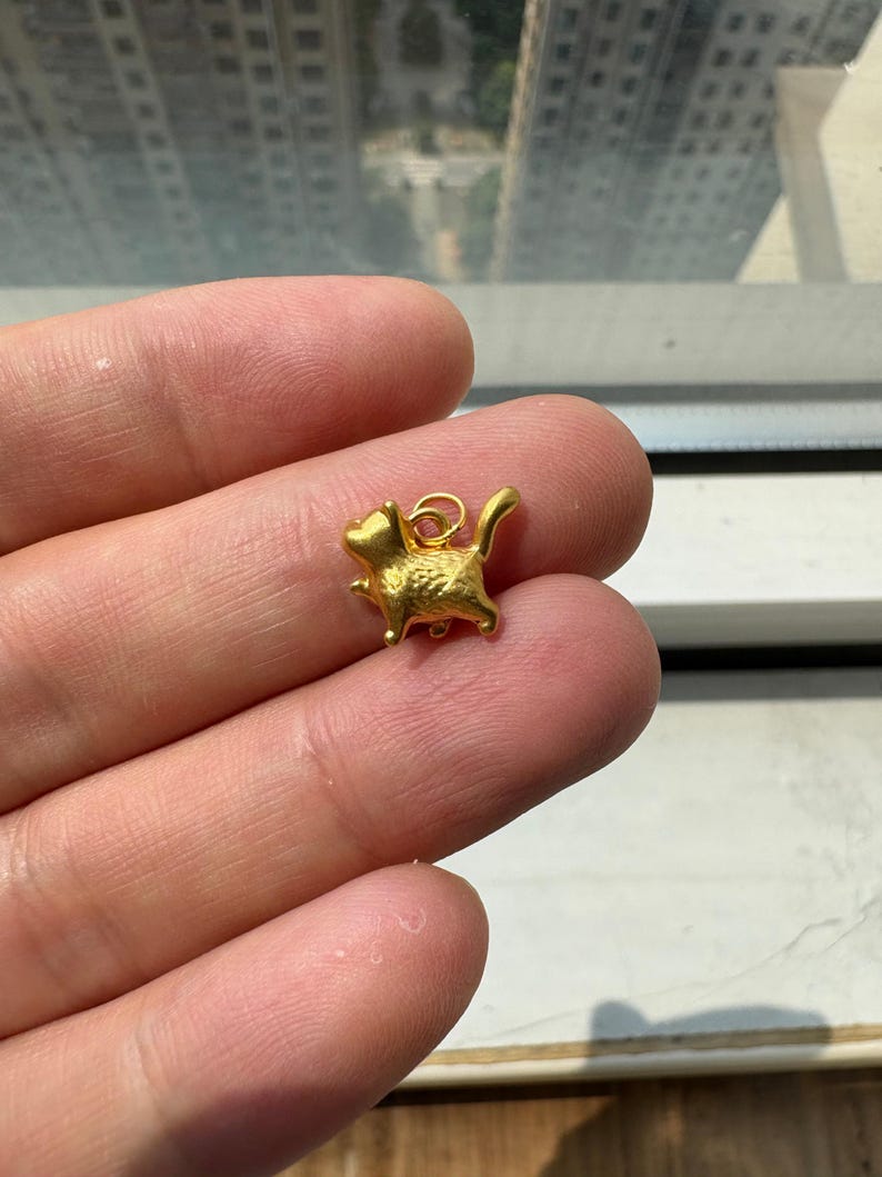 May include: A small, gold-colored metal charm shaped like a stylized cat in a walking pose. The charm features a textured surface and includes a small circular jump ring attached to the top of the cat's back for hanging purposes.