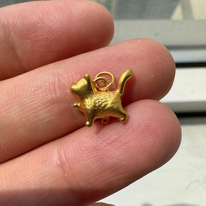 May include: A small, gold-colored metal charm shaped like a cat in a standing position. The cat figure has a textured surface and a small circular jump ring attached to its back for hanging. The charm is solid gold in color with a matte finish.