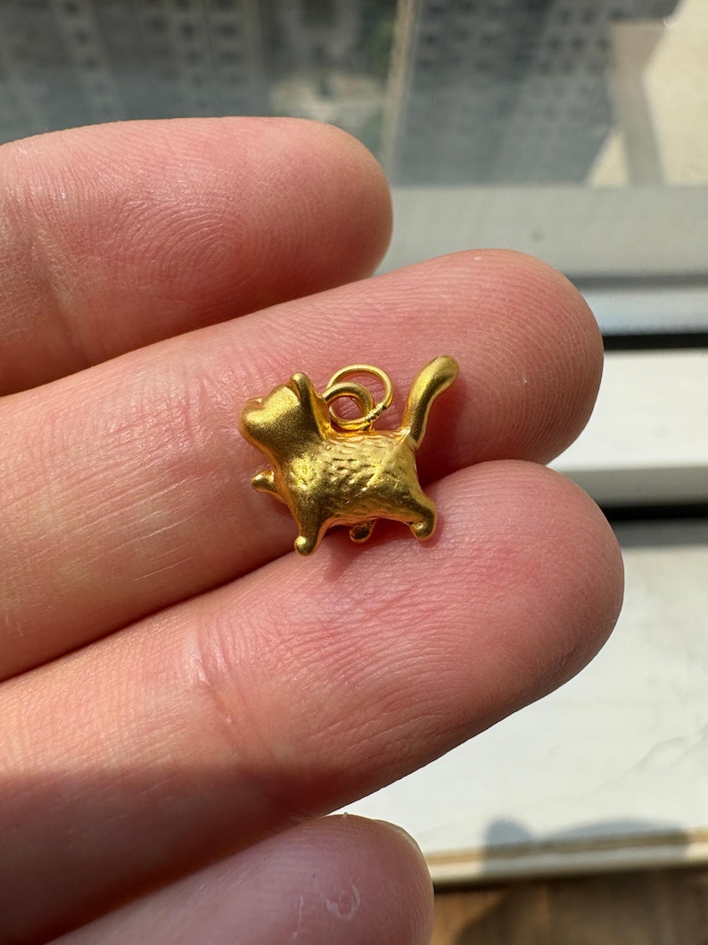 May include: A small, gold-colored metal charm shaped like a cat in a standing position. The cat figure has a textured surface and a small circular jump ring attached to its back for hanging. The charm is solid gold in color with a matte finish.