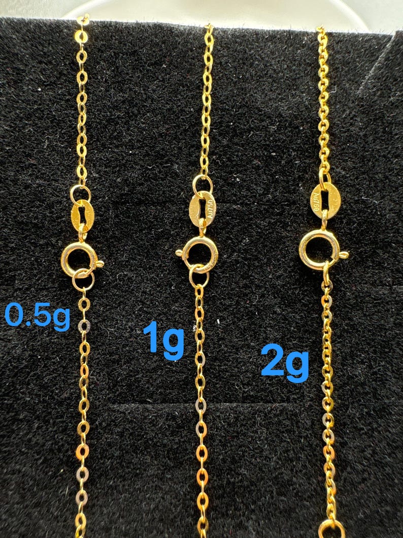 May include: Three gold chains with spring ring clasps, each featuring a small metal tag stamped with AU750. The chains are labeled with blue text indicating weights of 0.5g, 1g, and 2g, representing the specific mass of each individual gold chain.