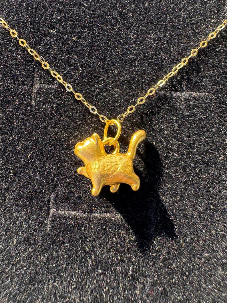 May include: A gold-coloured necklace with a cat-shaped pendant. The pendant shows a detailed cat with paws and a tail. The fine gold chain hangs against a dark background. The pendant is approximately 2 cm in length.