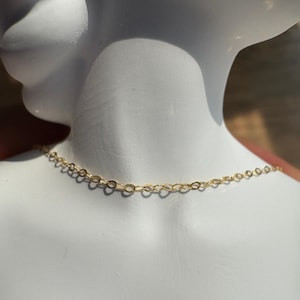 May include: A delicate gold chain necklace featuring small, uniform oval-shaped links. The chain is displayed resting against a smooth, white mannequin neck, highlighting the metallic yellow finish and the intricate, repeating pattern of the metal links.
