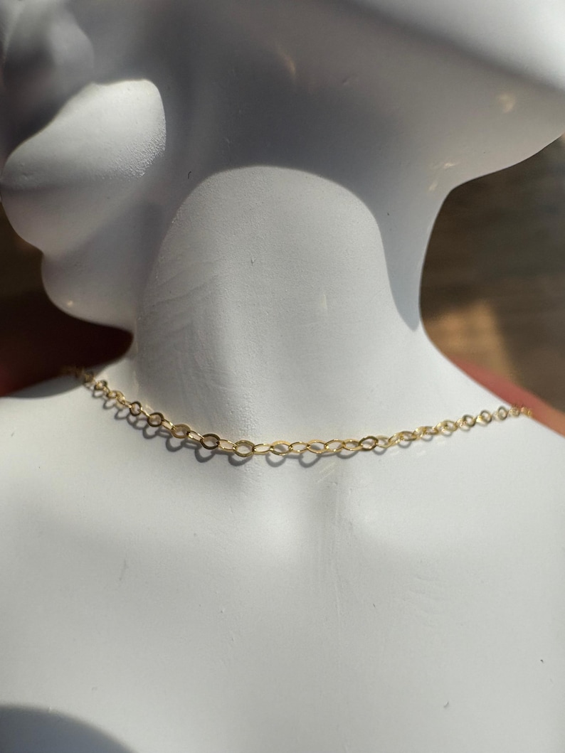 May include: A delicate gold chain necklace featuring small, uniform oval-shaped links. The chain is displayed resting against a smooth, white mannequin neck, highlighting the metallic yellow finish and the intricate, repeating pattern of the metal links.