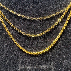 May include: A triple-layered gold chain necklace featuring three delicate, interconnected strands of polished gold cable links. The chains are arranged in a cascading, tiered formation, creating a layered look with uniform, rounded metal links.
