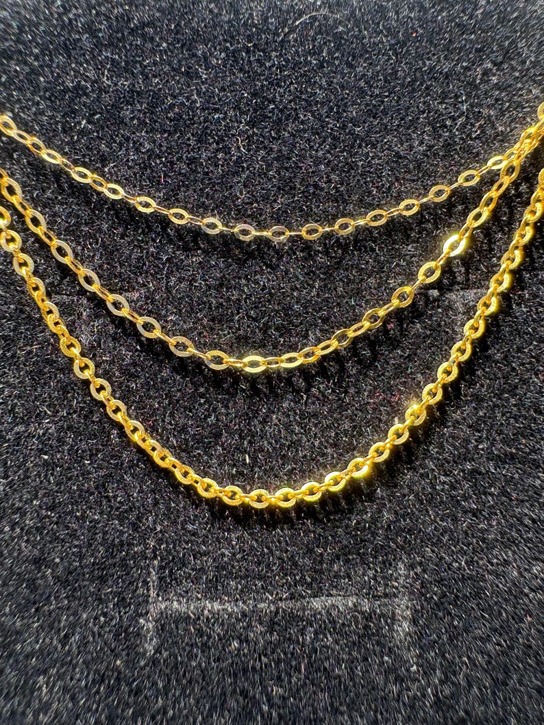 May include: A triple-layered gold chain necklace featuring three delicate, interconnected strands of polished gold cable links. The chains are arranged in a cascading, tiered formation, creating a layered look with uniform, rounded metal links.
