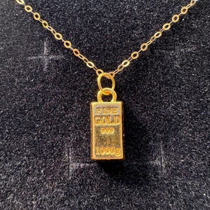 Könnte beinhalten: Eine goldfarbene Halskette mit einem rechteckigen Anhänger in Form eines Miniatur-Goldbarrens. Der Anhänger ist mit dem Text FINE GOLD, 999 und 1000g graviert. Er hängt an einer zierlichen goldfarbenen Kette mit kleinen, feinen Gliedern.
