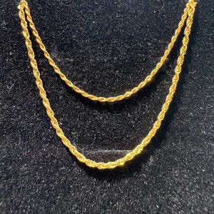 May include: A gold-toned rope chain necklace featuring a twisted, spiral link design. The chain is displayed in a curved, draped position, showcasing the metallic texture and reflective quality of the gold-colored links against a dark surface.