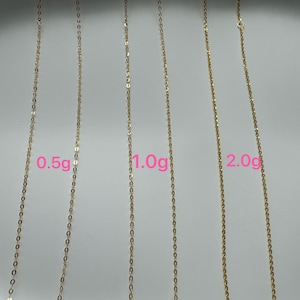May include: Three gold-colored metal chains of varying thicknesses displayed vertically. The chains are labeled with pink text indicating weights of 0.5g, 1.0g, and 2.0g respectively, showing the relative scale and thickness of each individual jewelry chain.
