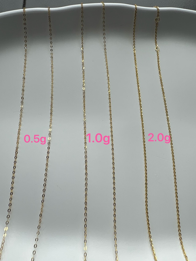 May include: Three gold-colored metal chains of varying thicknesses displayed vertically. The chains are labeled with pink text indicating weights of 0.5g, 1.0g, and 2.0g respectively, showing the relative scale and thickness of each individual jewelry chain.