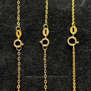 May include: Three gold chains featuring delicate oval links and circular spring ring clasps. Each chain includes a small oval tag stamped with the text AU750, indicating the gold purity. The chains are arranged vertically in a parallel formation.