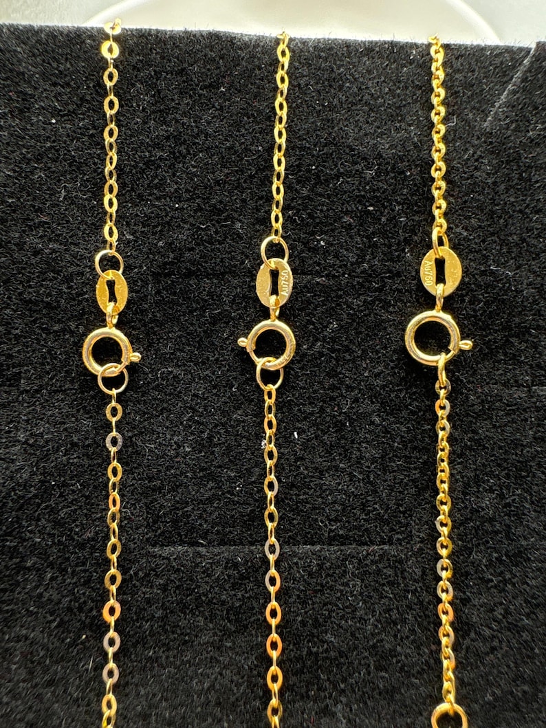 May include: Three gold chains featuring delicate oval links and circular spring ring clasps. Each chain includes a small oval tag stamped with the text AU750, indicating the gold purity. The chains are arranged vertically in a parallel formation.