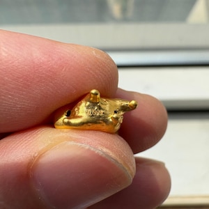May include: A small, textured gold charm featuring a rounded, organic shape with several protruding points. The surface of the gold piece is stamped with the text AU999, indicating its composition. The object has a bright, metallic yellow finish.