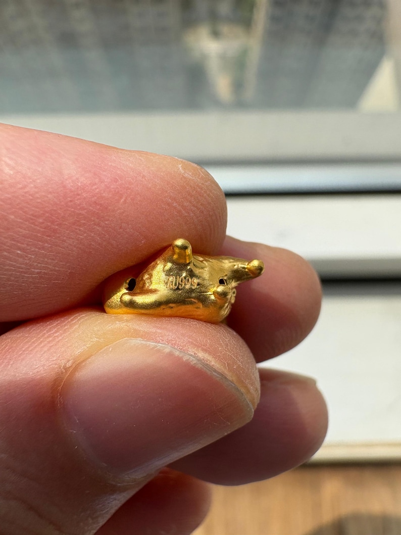 May include: A small, textured gold charm featuring a rounded, organic shape with several protruding points. The surface of the gold piece is stamped with the text AU999, indicating its composition. The object has a bright, metallic yellow finish.