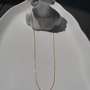 May include: A long, thin gold chain necklace with a delicate, reflective link design. The chain is draped over a white, smooth, sculptural bust display, showcasing its slender profile and metallic finish against the neutral, matte white surface.