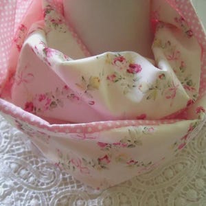 Könnte beinhalten: Ein rosa-weißer Infinity-Schal mit Blumenmuster und gepunktetem Besatz. Der Schal hat ein zartes Rosenmuster und ist aus weichem Stoff gefertigt. Der Schal ist über einer weißen Oberfläche mit Spitzendesign drapiert.