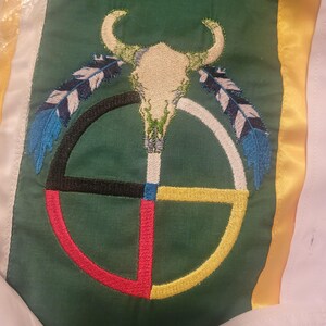 May include: A green fabric panel featuring an embroidered white bull skull centred above a circular medicine wheel design. The wheel is divided into black, white, red, and yellow quadrants. Two blue and white feathers flank the skull.