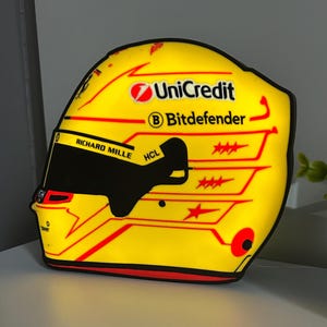 Könnte beinhalten: Ein gelber Rennhelm mit roten und schwarzen Akzenten. Der Helm zeigt die Logos von UniCredit und Bitdefender sowie die Texte "RICHARD MILLE" und "HCL".