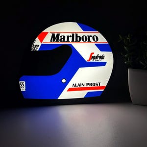 Peut inclure: Casque décoratif avec le logo Marlboro et le nom d'Alain Prost. Le design du casque intègre des couleurs rouge, blanc et bleu. Le casque est éclairé et placé sur un fond sombre.