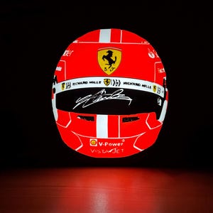 Peut inclure: Casque de course rouge et blanc avec le logo Ferrari et la signature d'un pilote. Le casque présente le logo V-Power VistaJet et la marque Richard Mille. Le fond est noir.