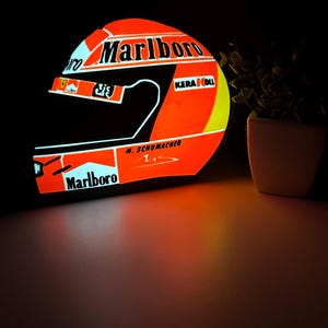 Könnte beinhalten: Ein leuchtend orange-schwarzer Rennhelm mit dem Marlboro-Logo und dem Namen M. Schumacher. Der Helm hat einen gelben Streifen und die Nummer 1. Das Bild ist vor einem dunklen Hintergrund.