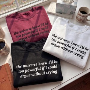 Può includere: Tre felpe girocollo bordeaux, nere e bianche, ciascuna ricamata con la frase "the universe knew I'd be too powerful if I could argue without crying". Le felpe sono esposte su un davanzale.